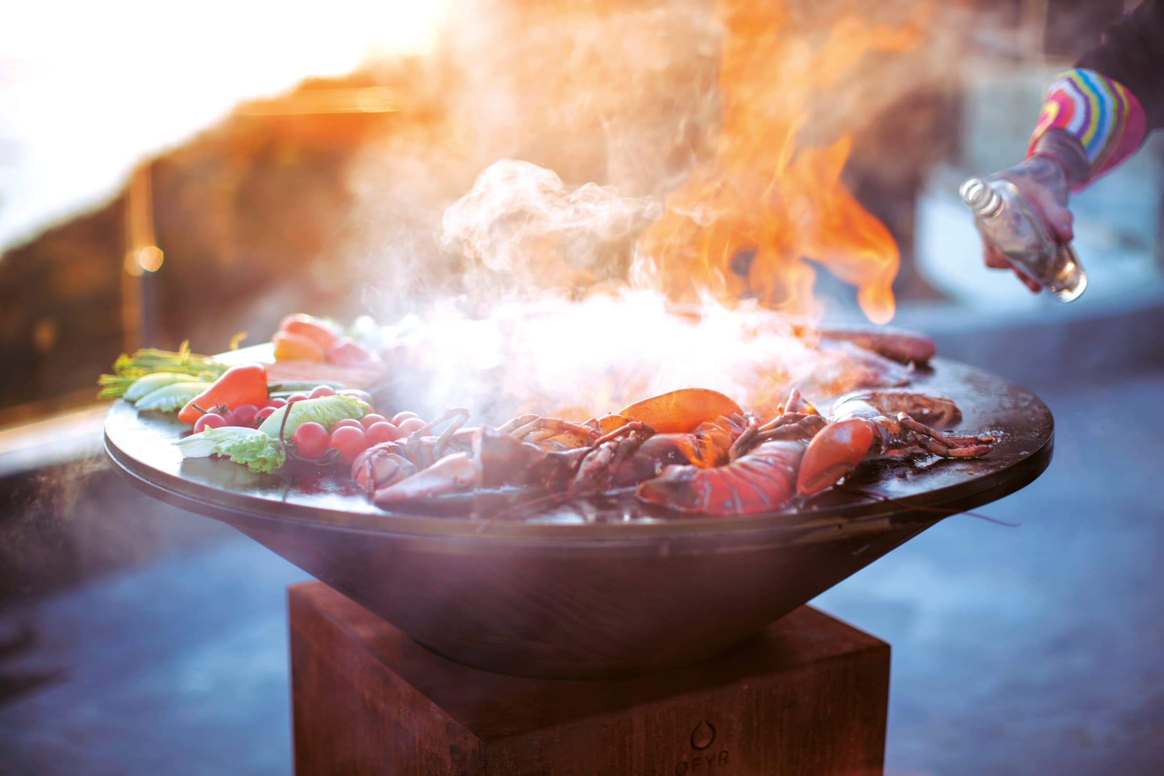 Plancha OFYR en pleine cuisson : fruits de mer, légumes et flammes au coucher du soleil