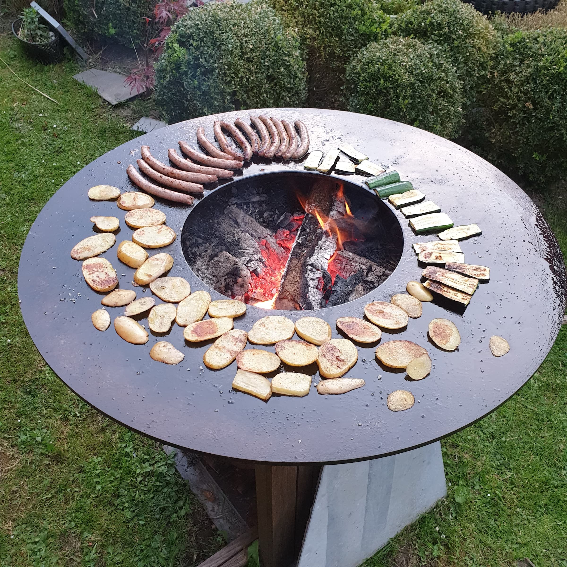 Vue de dessus d'un barbecue OFYR : saucisses, pommes de terre et courgettes grillées autour du foyer