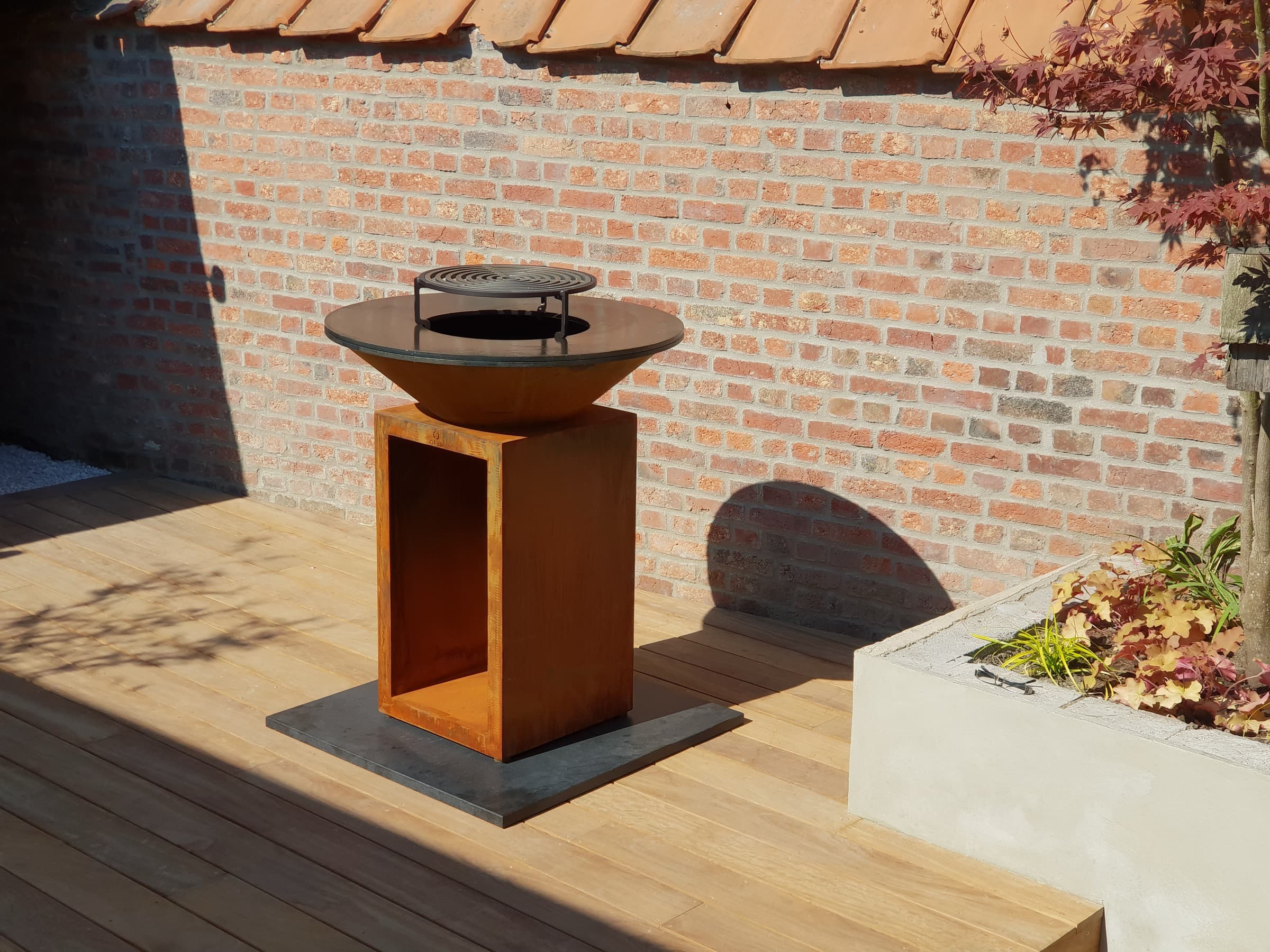 Barbecue OFYR en acier corten sur une terrasse en bois, contre un mur en briques